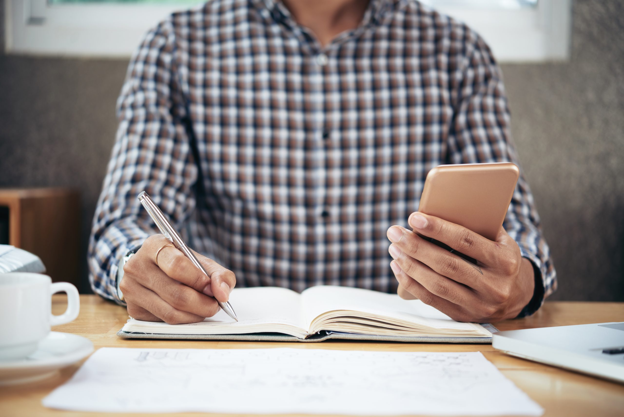 Crop shot of unrecognizable man sitting at office table, looking at smartphone and taking notes in register book